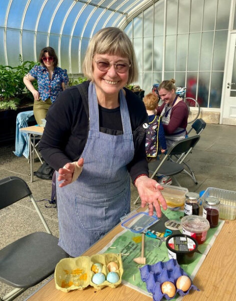 Image of smiling volunteer from glasgow flower garden wearing a blue apron, at a messy table in the Kibble Palace that is covered in egg-dying supplies like cups of dyes, in the Kibble Palace
